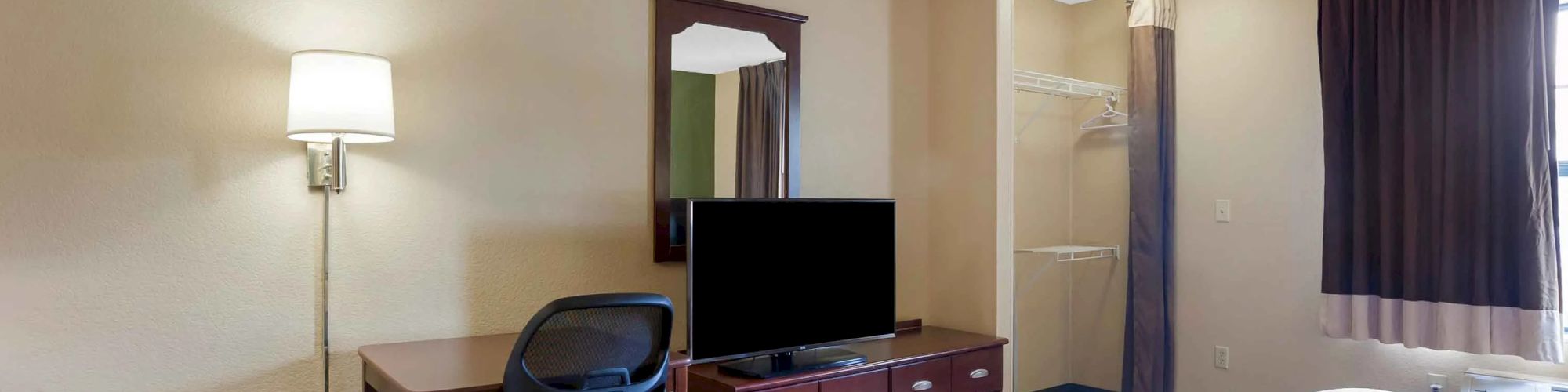 The image shows a hotel room with a bed, desk, chair, lamp, TV, dresser, mirror, and an open closet in the background.