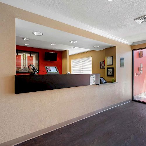 The image shows a hotel reception area with a brown front desk, a computer monitor, wall art, and a glass entrance door.