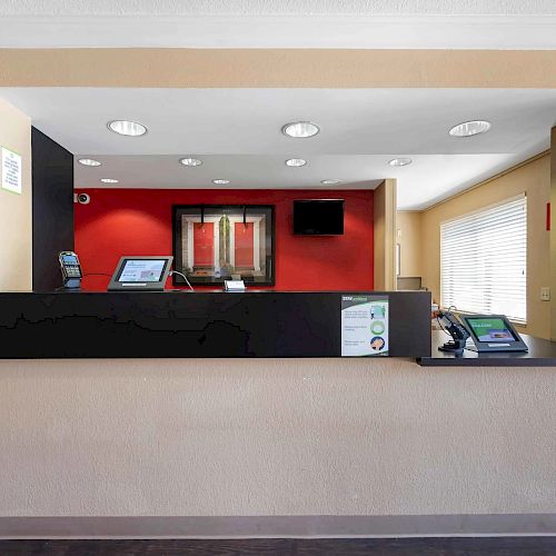 The image shows a hotel reception desk area with a computer, documents, and a small plant on the counter, featuring a red accent wall behind.