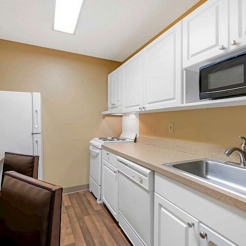 The image shows a kitchen with white cabinets, a sink, a microwave, a refrigerator, a stove, a counter, and two chairs.