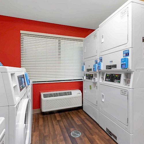 The image shows a laundry room with multiple white washing machines and dryers, a red wall, wooden floor, and a window with blinds.