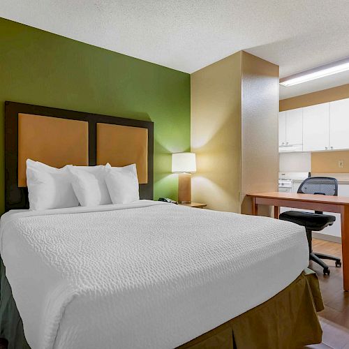 A hotel room with a bed, bedside table, lamp, desk, chair, and a kitchenette area. The walls are green and beige.