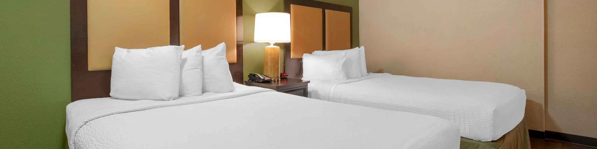The image shows a hotel room with two neatly made beds, a nightstand with a lamp between them, green and beige walls, and wooden flooring.