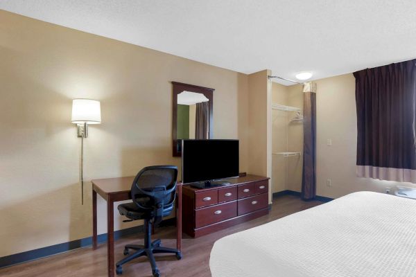 A hotel room with a bed, desk and chair, wall-mounted lamp, dresser with TV, mirror, open closet with hangers, curtains, and wooden floor.