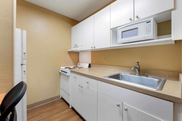 This image shows a compact kitchen with white cabinets, a microwave, sink, stovetop, and fridge, all set against beige walls and wooden flooring.