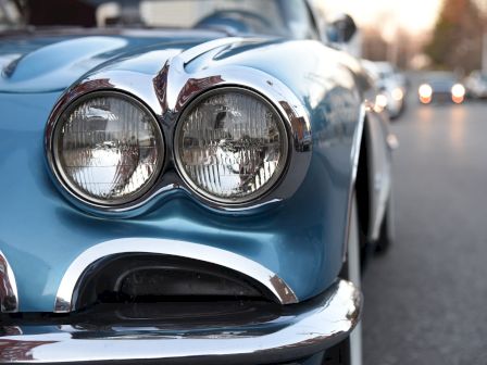 The image shows a close-up of the front end of a blue classic car with round headlights, parked on a street lined with other cars.