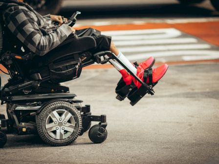 A person in a black and grey outfit sitting in a power wheelchair, wearing bright red shoes, and using a phone on a city street.