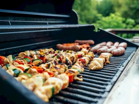 A barbecue grill with skewers of vegetables and meats, alongside sausages, set in an outdoor setting with greenery in the background.