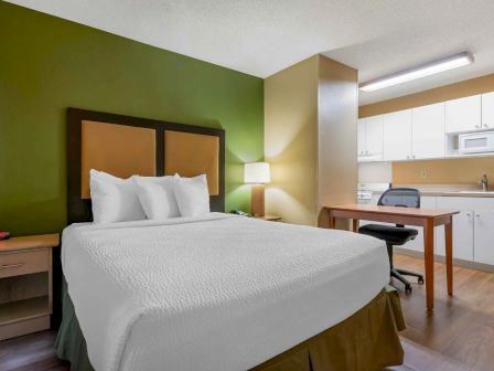 The image shows a hotel room with a bed, nightstands, lamp, desk, chair, and a kitchenette in the background.