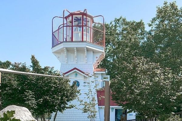 This image shows a quaint white lighthouse with red accents surrounded by trees and rocks on a clear day, always ending the sentence.