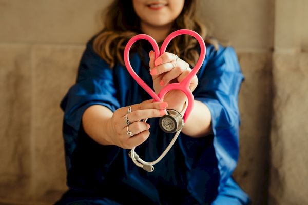A person in a blue outfit, possibly a medical professional, holding a pink stethoscope shaped like a heart in front of them.
