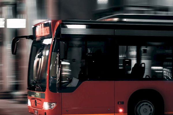 A red public bus is captured moving quickly through a city street at night, with urban buildings blurred in the background, suggesting speed.