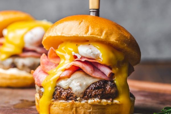 A juicy burger with melted cheese, ham, and a sauce, topped with a knife, sits on a wooden board, with another burger slightly blurred in the background.